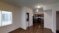 This single-floor space features an open-concept layout that connects the living area to the kitchen, creating a cohesive and functional flow. The dark hardwood-style flooring runs throughout, providing a warm contrast to the bright white walls and ceiling.

The kitchen is equipped with medium-toned wood cabinetry offering ample storage, dark stone-look countertops, and black appliances including a range, microwave, and refrigerator. Flush-mount ceiling light fixtures illuminate the space with adequate overhead lighting.

A dedicated laundry closet is conveniently tucked behind a paneled door, housing a stacked washer and dryer unit — a practical in-unit feature. The open living area offers a clean, neutral canvas with white walls and generous floor space. White baseboards frame the perimeter of the rooms, adding a clean, finished look. Overall, the space presents a straightforward, functional design with durable finishes suited for everyday living.