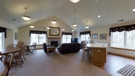This two-story space is highlighted by an expansive open-plan main living area defined by soaring vaulted ceilings that lend an airy, generous sense of volume throughout. A series of elegant pendant lights hang at varying heights from the ceiling, providing warm, layered illumination that complements the recessed lighting above. A stone fireplace serves as a natural focal point along one wall, flanked by windows dressed with dark floral valances that add a traditional touch to the interior. The living area is furnished with dark leather reclining sofas and armchairs arranged around a central coffee table, creating a comfortable and inviting gathering space. A flat-screen television is positioned near the fireplace, enhancing the room's functionality. A decorative gold-framed mirror adorns the wall, adding depth and elegance to the space. Adjacent to the living area is a counter-height bar or dining space with wooden stools, seamlessly integrating a casual dining or social zone into the open floor plan. The flooring transitions from plush neutral carpet in the main seating area to hard flooring near the counter space. Framed artwork on the walls adds character and a refined finishing touch to the overall interior.