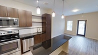 This single-floor 1-bedroom, 1-bathroom residence is thoughtfully designed to maximize both functionality and style. The open-concept kitchen flows seamlessly into the living area, creating a cohesive and inviting space. The kitchen is a standout feature, showcasing rich dark wood cabinetry, sleek dark granite countertops, and a peninsula that doubles as a breakfast bar — ideal for casual dining or entertaining. Stainless steel appliances, including a dishwasher, are complemented by a stainless undermount sink and modern pendant lighting that adds warmth and visual interest overhead. A tall pantry-style cabinet provides generous storage, while open shelving adds a contemporary touch. Mosaic tile backsplash detailing adds a refined accent to the kitchen walls. Light-toned hardwood-style flooring extends throughout, creating a clean and consistent aesthetic. The adjacent living area benefits from natural light streaming through a window, lending an airy quality to the space. Neutral wall tones throughout keep the interior feeling bright and versatile. The overall ambiance is modern and polished, offering a comfortable and well-designed environment suited for everyday living.