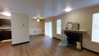 This single-floor residence offers a welcoming and well-appointed living space defined by rich, dark hardwood flooring that runs throughout the main area, lending warmth and a sense of continuity. The living room serves as the heart of the home, highlighted by a classic fireplace with a dark surround and a white mantel, providing a cozy focal point. Crown molding frames the textured ceiling, adding a refined architectural detail that complements the neutral, warm-toned walls. A ceiling fan with a light fixture is centrally positioned, ensuring comfort and airflow throughout the space. Large sliding glass doors open to the outside, allowing generous amounts of natural light to pour in and creating a seamless connection between the interior and the outdoors. An adjacent window further enhances the bright, airy atmosphere. A secondary area, visible beyond the main living space, features wainscoting along the lower walls and an additional ceiling fan, suggesting a versatile dining or flex room. A pet door is also integrated into the wall, adding a practical touch. The overall ambiance is comfortable, bright, and inviting, blending classic design elements with functional, everyday living in mind.