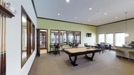 This single-floor amenity space is thoughtfully designed to serve as a social and recreational hub. The main game room is anchored by a full-size pool table positioned at the center of the carpeted floor, providing ample space for play and movement. Soft green accent walls contrast with crisp white trim and dark wood crown molding, creating a welcoming and energetic atmosphere. Recessed lighting throughout the ceiling ensures bright, even illumination across the entire area. Along one wall, a row of wood-framed windows overlooks an adjacent space outfitted with high-top bar tables and stools, offering a casual spot for conversation or spectating. A wall-mounted flat-screen television adds an entertainment focal point to the room. Additional lounge seating is arranged toward the far end of the space, providing a relaxed area to unwind. A glass-paned door leads to what appears to be a fitness room, making this a comprehensive recreational facility. The combination of game, lounge, and fitness areas within a single cohesive layout makes this space well-suited for community gatherings and everyday leisure activities.