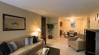 This single-floor residence offers a thoughtfully designed open-concept layout that seamlessly connects the main living areas. The living room serves as the heart of the home, featuring plush wall-to-wall carpeting, neutral beige walls, and a textured ceiling that adds subtle character. Warm ambient lighting from a classic table lamp creates a cozy, inviting atmosphere, complemented by a decorative wall mirror that adds depth to the space. Built-in shelving provides practical storage and display opportunities. The living area flows naturally into the dining space, which is brightened by a ceiling fan with integrated lighting. Elegant dining chairs surround the table, making it an ideal setting for meals and gatherings. Adjacent to the dining area, the kitchen is visible in the background, featuring dark wood cabinetry that provides a rich contrast to the lighter tones throughout the home. Decorative touches, including a potted indoor plant and wall art, add personality and a sense of warmth throughout. The open layout ensures easy movement between spaces, making this residence as functional as it is comfortable. The consistent flooring and color palette throughout tie the space together into a harmonious and welcoming environment.