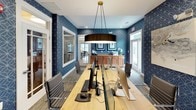 Located at 101 Chesapeake Ridge, this single-floor business center offers a well-appointed workspace designed to support productivity and comfort. The room makes a strong visual impression with its bold navy blue geometric-patterned wallcovering that extends throughout, creating a cohesive and energetic atmosphere balanced by crisp white trim and a light ceiling.

At the heart of the space is a long, natural wood communal workstation accommodating multiple users, outfitted with iMac and PC computers, keyboards, and mice. Sleek, modern rolling chairs in silver and gray provide ergonomic seating for each workstation. A large cylindrical drum pendant light hangs centrally above the table, anchoring the room and providing warm, focused illumination alongside recessed ceiling lights.

A wall-mounted flat-screen television adds multimedia functionality, while a warm wood credenza along one wall offers additional storage and display space, accented with decorative blue and white accessories. Curated artwork, including an abstract canvas piece and a framed feather print, add personality to the space. Large windows fitted with wooden blinds invite natural light, and glass-paneled interior windows and doors maintain a sense of openness and visual connectivity between areas. Neutral gray carpeting grounds the room with a clean, professional finish.
