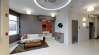 This single-floor commercial space serves as the office of Blackstream | Christie's International Real Estate, showcasing a refined and contemporary design throughout. The reception and lounge area is immediately striking, featuring a glossy white floor that reflects light and enhances the sense of openness. A distinctive circular LED light fixture is recessed into the ceiling, creating a dramatic focal point overhead and contributing to the space's polished, modern aesthetic. Pendant lights provide additional warm illumination throughout.

The lounge area is furnished with a clean-lined white sofa accented with neutral throw pillows, positioned around a wooden and glass coffee table set atop a rich, dark shag area rug. A sculptural metallic console table against the far wall, paired with decorative black wall art and elegant candle holders, adds artistic character to the space.

A glazed sliding door with high-gloss red panels leads to a private inner office, visible through the glass and featuring a wall-mounted television and a sleek white workstation. The bold red accent wall inside creates a vivid contrast against the predominantly grey and white palette of the reception area, lending the overall space a dynamic and professional energy.