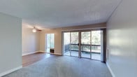 This single-floor residence offers a thoughtfully designed layout that balances comfort and functionality. The living area is generously proportioned, featuring plush carpeting, neutral-toned walls, and a textured ceiling. A ceiling fan provides year-round climate comfort, while large sliding glass doors open directly onto a private balcony that overlooks lush, mature greenery — allowing abundant natural light to fill the space. A wall-mounted heating unit adds to the room's practical amenities.

The kitchen is designed in a classic galley configuration, maximizing efficiency with white shaker-style cabinetry, dark countertops, and wood-look flooring. It is equipped with black and stainless steel appliances, including a refrigerator, electric range with overhead ventilation, and dishwasher, offering ample storage and counter space throughout.

The bedroom is a calm, carpeted retreat with a large window that draws in natural light, complemented by a baseboard heater and a textured ceiling. The bathroom features a full tub-and-shower combination with clean white surround, chrome fixtures, a curved shower rod, and grab bars, reflecting a clean and accessible design.

Overall, the space presents a clean, move-in ready environment with a cohesive neutral palette, practical finishes, and inviting outdoor connectivity.