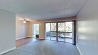 This single-floor residence offers a thoughtfully designed layout that balances comfort and functionality. The living area is generously proportioned, featuring plush carpeting, neutral-toned walls, and a textured ceiling. A ceiling fan provides year-round climate comfort, while large sliding glass doors open directly onto a private balcony that overlooks lush, mature greenery — allowing abundant natural light to fill the space. A wall-mounted heating unit adds to the room's practical amenities.

The kitchen is designed in a classic galley configuration, maximizing efficiency with white shaker-style cabinetry, dark countertops, and wood-look flooring. It is equipped with black and stainless steel appliances, including a refrigerator, electric range with overhead ventilation, and dishwasher, offering ample storage and counter space throughout.

The bedroom is a calm, carpeted retreat with a large window that draws in natural light, complemented by a baseboard heater and a textured ceiling. The bathroom features a full tub-and-shower combination with clean white surround, chrome fixtures, a curved shower rod, and grab bars, reflecting a clean and accessible design.

Overall, the space presents a clean, move-in ready environment with a cohesive neutral palette, practical finishes, and inviting outdoor connectivity.