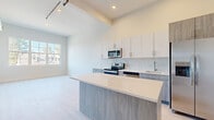 This thoughtfully designed single-floor residence showcases a clean, contemporary aesthetic throughout. The heart of the unit is an open-concept kitchen that flows seamlessly into the living area. The kitchen is appointed with two-tone cabinetry — crisp white upper cabinets paired with warm wood-textured lower cabinets — complemented by a marble-style backsplash and bright quartz countertops. A spacious center island provides additional prep space and informal seating. Stainless steel appliances, including a French door refrigerator, electric range, built-in microwave, and dishwasher, complete the well-equipped culinary space. Elevated ceilings and recessed lighting add a sense of openness and volume to the kitchen and living area, while industrial-style track lighting introduces an urban edge. Light-toned hardwood flooring runs throughout the main living spaces, amplifying the unit's bright and airy atmosphere. A generously sized bedroom benefits from a wide bank of windows that invite abundant natural light and outdoor views. The bathroom features marble-style tile flooring and a wood-grain vanity with a clean white countertop, maintaining the modern and cohesive design language found throughout the entire space.