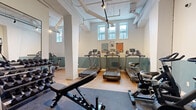 Located at 87 Mann Ave in Ottawa's Sandy Hill neighbourhood, this fitness facility is thoughtfully designed to maximize function within a compact footprint. The space is organized into two distinct zones that together provide a comprehensive workout environment. The main cardio and strength area features multiple treadmills, stationary bikes, and an elliptical trainer arranged along light-washed walls accented with a two-tone teal and white colour scheme. An Inspire cable machine and a full rack of dumbbells serve the strength training corner, complemented by a large wall mirror that enhances the sense of space and supports form monitoring. A secondary area hosts a row of spin bikes and an adjustable weight bench, ideal for group or solo cycling sessions. Light wood flooring transitions to durable rubber mat zones beneath the equipment, balancing aesthetics with practicality. Exposed ductwork and track lighting on the ceiling lend the space an industrial character, while a row of windows along one wall introduces natural light. A striking illustrated mural depicting cyclists in an outdoor mountain setting adds a vibrant, motivating focal point to the room, reinforcing an active and energetic atmosphere throughout.