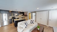 This single-floor space showcases an open-concept layout that seamlessly connects the living and kitchen areas, creating a cohesive and functional environment. The living area is anchored by a comfortable neutral-toned sofa accented with patterned throw pillows, paired with a dark wood coffee table that adds contrast to the warm hardwood-style flooring running throughout the space.

The kitchen is a standout feature, designed with dark espresso cabinetry that extends to the upper and lower sections, providing ample storage. Light granite countertops offer a striking contrast against the dark cabinetry, while stainless steel appliances — including a range, microwave, and refrigerator — complete the modern aesthetic. Pendant lighting hangs above the breakfast bar, which accommodates seating and doubles as a casual dining space.

Neutral grey walls and a textured ceiling contribute to a clean, contemporary ambiance. A set of white panel doors leads to additional rooms, including what appears to be a bathroom and a closet or secondary room, suggesting a practical and well-organized floor plan throughout the unit.