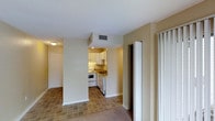 This one-bedroom, one-bathroom unit is thoughtfully laid out across a single floor, making efficient use of its space. The open living area flows naturally toward a semi-enclosed kitchen, which is defined by a partial dividing wall that creates a sense of separation while maintaining an open feel. The kitchen is equipped with white cabinetry, a white range, a dishwasher, and a sink set beneath under-cabinet lighting, all complemented by tile flooring that extends throughout the kitchen and living areas. The neutral, warm-toned walls and light-colored ceiling provide a clean, bright backdrop throughout the unit. A ceiling-mounted HVAC vent ensures year-round climate control. The living space transitions to carpeted areas, adding a layer of comfort underfoot. A sliding door closet provides convenient storage, while additional doorways lead to the bedroom and bathroom, each offering privacy and functionality. The textured ceiling and flush-mount lighting fixture contribute to the unit's straightforward, utilitarian aesthetic. Overall, the space presents a practical and cohesive layout well-suited for comfortable everyday living.