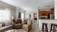 This single-floor residence is designed with an open, interconnected layout that promotes a comfortable and functional flow between spaces. The living room serves as the heart of the home, featuring plush carpeting, crown molding, and multiple windows dressed with horizontal blinds that allow generous natural light. A neutral color palette of warm grays and beige tones creates a cohesive, inviting atmosphere throughout.

The dining area is positioned adjacent to the living room and benefits from a chandelier overhead and sliding glass doors that provide outdoor views and additional light. The kitchen is well-equipped with warm wood cabinetry, granite-style countertops, stainless steel appliances including a dishwasher and microwave, and hardwood-style flooring. A pass-through window and breakfast bar connect the kitchen to the main living area.

The bedroom is a spacious retreat with carpeted flooring, a ceiling fan, and a wall of mirrored sliding closet doors that enhance the sense of space. The bathroom features hardwood-style flooring, a tub-shower combination, and granite-topped vanity. The overall ambiance is warm, classic, and well-maintained.