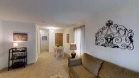 This single-floor residential space offers a welcoming and functional layout designed for comfortable everyday living. The open-plan living and dining area serves as the heart of the home, where soft carpeted flooring and neutral white walls create a warm, understated ambiance throughout. The living area is cozy and inviting, highlighted by a large curtained window that allows natural light to fill the space, complemented by decorative wall art that adds a touch of character. Flowing seamlessly from the living area, the dining space accommodates a small table and chairs, making it ideal for casual meals. A ceiling light fixture provides additional illumination to the dining area, while a wooden door nearby hints at additional storage or closet space. A hallway extends further into the unit, suggesting access to additional rooms beyond — likely bedrooms and a bathroom — all finished with the same consistent carpeting and clean wall tones. The overall aesthetic is practical and neutral, offering a comfortable, move-in-ready environment that serves as an excellent blank canvas for personalization.