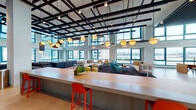 This generously scaled, single-floor amenity space is defined by its dramatic double-height ceilings and an extensive wraparound curtain wall of teal-framed windows that frame sweeping views of a waterway and urban skyline beyond. Abundant natural light floods the interior throughout the day, creating a bright, energizing atmosphere.

The space is organized into several distinct functional zones. A central lounge area features a bold grid of exposed black steel ceiling beams from which a curated collection of globe and pendant light fixtures are suspended, adding visual interest overhead. Multiple seating arrangements—ranging from large sectional sofas and modular lounge chairs to teal accent chairs and colorful ottomans—encourage both social gathering and quiet relaxation.

A long, light-wood bar counter with vibrant red barstools serves as a casual workspace or social hub, while a separate wet bar area showcases a striking textured teal tile backsplash with pendant lighting above. A dedicated quiet workspace area offers a streamlined desk with waterfront views. A serene meditation or wellness nook features built-in tiered seating with cushions and large windows overlooking the waterway. Additional seating vignettes with crisp white armchairs provide quieter retreat spaces throughout.