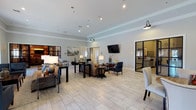This single-floor clubroom is thoughtfully designed to accommodate a variety of social functions, offering multiple distinct zones across a generous, open layout. The main gathering area serves as the centerpiece of the space, furnished with plush sofas and accent chairs arranged around a central coffee table, creating an inviting lounge environment ideal for casual conversation and relaxation. A wall-mounted television anchors one end of the lounge area, enhancing its functionality for group viewing.

Adjacent to the lounge, a dedicated dining area features a substantial table with seating for multiple guests, making it well-suited for community events and organized gatherings. The space also includes a separate dining nook and what appears to be a kitchenette or catering area, supporting food and beverage service during events.

Light-toned tile flooring runs throughout, lending a clean and cohesive aesthetic across all zones. Recessed lighting and stylish gooseneck wall sconces provide warm, even illumination, complementing the neutral color palette of soft grays and whites. Crown molding adds a refined architectural detail, while glass-panel partitions define areas without compromising the open, airy feel of the overall space.