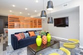 This single-floor communal space is designed with a bold, contemporary aesthetic that balances energetic color with clean, modern architecture. The lounge area anchors one end of the space, featuring a deep navy sofa accented with colorful throw pillows, a sleek wood coffee table adorned with vivid green vases, and a pair of striking yellow wire-frame chairs. A cluster of matte black pendant lights hangs overhead, while large windows fitted with white blinds flood the area with natural light and offer views to the exterior. A wall-mounted flat-screen television and a vibrant orange slatted room divider add character and definition to the space.

The kitchen area is outfitted with a large island featuring a crisp white countertop and grey cabinetry, paired with orange-topped bar stools for casual seating. Dark, textured backsplash tiles and dark upper cabinetry contrast beautifully with stainless steel appliances, including a full-size refrigerator. A linear pendant light adds an industrial touch overhead.

The dining zone features a large, rustic-style reclaimed wood table surrounded by upholstered chairs, positioned near a window for natural light. Bold, large-scale colorful artworks line the walls, adding a gallery-like energy to the space. Throughout, a multi-tone striped hardwood floor ties all zones together cohesively.