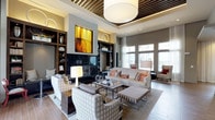 This thoughtfully designed lobby serves as a welcoming communal space that combines sophisticated aesthetics with functional comfort. The lounge area anchors the space with a curated seating arrangement, including a neutral-toned sofa accented by vibrant patterned throw pillows, upholstered armchairs, and a striped ottoman gathered around a sleek rectangular coffee table. A striking yellow abstract painting commands attention above a modern fireplace framed by dark built-in shelving units, which display decorative vases and curated accessories, adding depth and visual interest to the room. The ceiling features an eye-catching design with slatted wood panels and recessed lighting alongside a large, contemporary drum pendant light that anchors the space from above. Floor-to-ceiling windows and glass doors along one wall flood the interior with natural light, offering views to the exterior and creating an airy, open ambiance. Warm wood-toned flooring runs throughout, complementing the neutral gray and taupe color palette. The overall atmosphere is polished and inviting, projecting a sense of refined comfort appropriate for a welcoming lobby environment.