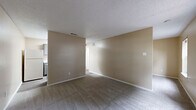 This one-bedroom, one-bathroom unit offers a thoughtfully arranged single-floor layout that maximizes its compact footprint. The living area is open and functional, featuring neutral beige walls, soft carpeted flooring, a textured ceiling, and two windows that invite natural light and views of the surrounding greenery. The space provides a clean, versatile canvas well-suited for a variety of furniture arrangements.

The kitchen is designed in a galley style, running along two parallel walls to make efficient use of space. It features gray cabinetry with marble-patterned countertops, wood-look vinyl flooring, a stainless steel sink, an electric range with overhead ventilation, and a full-size refrigerator. Upper and lower cabinets provide ample storage for everyday needs.

The bedroom is a cozy, carpeted room finished in warm neutral tones, offering a quiet retreat with access to a closet for storage. White trim and doors throughout the unit provide clean contrast against the warm wall colors.

A dedicated bathroom and an additional unclassified room round out the layout, contributing to the overall utility of the space. The consistent use of neutral tones, textured ceilings, and carpeted living areas gives the unit a cohesive, understated character throughout.