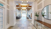 Located at 11115 Shadow Grove Cir in Charlotte, NC, this single-floor community space offers a sophisticated and well-appointed environment designed for both comfort and functionality. The grand entrance welcomes visitors with multicolored slate tile flooring, soaring double-height ceilings, ornate chandelier lighting, and warm wood-framed French doors that set an elegant tone throughout.

The primary living room is a showstopper, featuring dramatic two-story ceilings with richly stained wood coffered beams, a floor-to-ceiling stone fireplace, and expansive windows overlooking an outdoor pool area. Built-in display shelving frames a wall-mounted TV, while ample seating arrangements make this space ideal for gathering. A second, more intimate living area offers wainscoted walls, soft neutral tones, recessed lighting, and flexible seating with wall-mounted media.

The kitchen is thoughtfully designed with white shaker cabinetry, open floating shelves, a striking herringbone tile backsplash, stainless steel appliances, and the same slate tile flooring that flows throughout the main areas. The dedicated office features a built-in dual-workstation desk framed by floor-to-ceiling shelving with wainscot panel detailing, providing a professional and productive setting. Together, these spaces offer a refined and functional clubhouse experience.