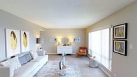 This single-floor space showcases a well-appointed living area designed with a clean, contemporary aesthetic. The room is finished with light hardwood-style flooring that contributes to an airy, open feel throughout. Neutral-toned walls provide a calm backdrop, complemented by a textured ceiling that adds subtle architectural interest.

A large sliding glass panel along one wall allows an abundance of natural light to flood the interior, creating a bright and welcoming atmosphere. The living area is thoughtfully arranged, anchored by a plush white sofa paired with a soft area rug. A sleek white credenza serves as a focal point along the far wall, flanked by matching table lamps and accented with decorative artwork and coastal-inspired décor pieces.

A pop of color is introduced through an orange accent chair, offering visual contrast against the predominantly neutral scheme. A sculptural coffee table centerpiece and a round side table further enhance the curated, modern styling of the space. Overall, the room balances functionality with refined design, making it both visually appealing and practically arranged.