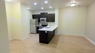 This single-floor space showcases a clean, contemporary design with an open-concept layout that promotes a sense of spaciousness and easy flow between areas. The kitchen serves as a central feature, equipped with dark espresso-toned cabinetry that provides a striking contrast against the crisp white countertops. A peninsula with an integrated sink extends into the living area, offering additional workspace and casual seating potential while reinforcing the open layout.

Stainless steel appliances, including a refrigerator, electric range, and microwave, lend a polished, modern aesthetic to the kitchen. Upper cabinetry provides ample storage, complemented by a tile backsplash that adds a subtle textural element. Recessed lighting throughout the ceiling ensures bright, even illumination across the space.

Light-toned hardwood-style flooring runs continuously through the kitchen and adjoining living area, enhancing the sense of openness. The neutral white walls further contribute to the airy, well-lit atmosphere. The overall design is minimal and functional, making effective use of the available footprint while maintaining a cohesive, contemporary visual identity.