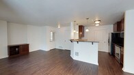 This single-floor space showcases an open-concept layout that seamlessly connects the living area and kitchen, creating a functional and airy environment. Dark hardwood-style flooring runs throughout, providing a rich contrast against the crisp white walls and textured ceiling.

The kitchen features a white peninsula with an overhang that accommodates bar seating, making it ideal for casual dining or entertaining. Rich dark wood cabinetry tops the peninsula, complemented by elegant pendant lights that hang above, adding a refined touch. The kitchen is further illuminated by a flush-mount ceiling fixture, ensuring ample lighting throughout.

The open living area is generously proportioned, offering flexibility in furniture arrangement. A window near the entryway allows natural light to filter into the space. The front entry door, framed in white, provides a clean and welcoming focal point. Additional doors visible in the space suggest access to further rooms or storage areas.

Overall, the space presents a clean, modern aesthetic with a neutral palette, warm flooring, and thoughtful lighting, creating a balanced and inviting atmosphere suited to everyday living.