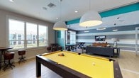 This lively recreational space is thoughtfully designed to encourage socializing and entertainment. At the heart of the room sits a striking pool table with a bold yellow felt surface, illuminated by two large, dome-shaped pendant lights that draw the eye and anchor the space. The design palette is energetic yet cohesive, with teal accent walls and horizontal grey and white striped walls adding a dynamic visual rhythm throughout.

Large windows along one wall flood the space with natural light, offering views of the surrounding outdoor landscape and creating an open, airy atmosphere. The light hardwood flooring complements the bright color scheme and ties the various areas of the room together seamlessly.

Beyond the pool table, the space opens into a multifunctional lounge and gaming area. Round gaming tables with high-backed chairs provide dedicated spots for card games or tabletop activities, while a ping-pong table offers additional recreational options. A comfortable seating arrangement featuring a plush grey sectional sofa, paired with a wall-mounted flat-screen television and a wooden media console, creates a relaxed lounge corner ideal for unwinding.

Overall, the space balances an energetic, playful aesthetic with comfortable, functional furnishings, making it an inviting destination for group activities and leisure.