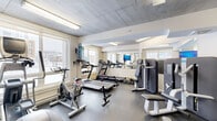 Located at 3400 Boul St-Elzéar O in Laval, Quebec, this fitness facility is a compact yet well-equipped gym designed to support a well-rounded workout routine. The space features an exposed concrete ceiling with flush-mounted fluorescent lighting, lending an industrial aesthetic that is both modern and functional. Dark rubber flooring throughout provides durability and safety underfoot.

Large windows along the perimeter allow considerable natural light to fill the room while offering views of the surrounding Laval neighbourhood. A full-length mirror on one wall visually expands the space and aids users during strength training exercises.

The gym is equipped with a variety of cardio machines, including SOLE-brand treadmills and spinning bikes, as well as a Precor elliptical. Strength training options include multiple selectorized resistance machines, a set of free weights and barbells, a stability ball, and a flat weight bench. Wall-mounted televisions are positioned for easy viewing during cardio sessions.

A split-unit air conditioning system ensures a comfortable temperature year-round. Additional amenities include a paper towel dispenser, a small utility shelf, and a waste bin to maintain cleanliness. The overall ambiance is clean, practical, and well-organized, making it a functional fitness amenity.