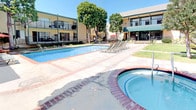 This outdoor amenity space centers around a generously sized swimming pool with crystal-clear turquoise water, complemented by a circular brick-rimmed spa positioned along the pool deck. The surrounding courtyard is finished with a combination of concrete and brick-accented paving, providing ample space for lounging and outdoor entertaining. Multiple chaise lounge chairs are arranged poolside, offering dedicated areas for sunbathing, while outdoor dining tables with chairs and a vibrant red patio umbrella create a comfortable setting for alfresco gatherings. Mature trees are strategically placed throughout the courtyard, offering natural shade and greenery that enhances the resort-like atmosphere. The complex itself features multi-story buildings with balconies and green-trimmed facades on one side and warm-toned stucco exteriors on the other, enclosing the courtyard to create a semi-private, sheltered environment. Stairways provide access between levels, and lush landscaping with shrubs and palms borders the perimeter. The overall ambiance is bright, open, and relaxed, well-suited for outdoor recreation and socializing under sunny skies.