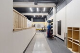 Located at 426 S Tejon St in Colorado Springs, CO, this fitness facility offers a thoughtfully designed single-floor layout optimized for a comprehensive workout experience. The space features an industrial-modern aesthetic, with exposed black-painted ceiling infrastructure, bright linear LED lighting, and a clean contrast of white and dark walls accented by bold yellow trim details.

The main workout area is divided into distinct functional zones. A cardio section houses multiple treadmills and a stationary bike, lined against a large wall mirror and complemented by a striking geometric mandala wall graphic. Nearby, a versatile open exercise zone features stability balls, medicine ball racks, and a wall-mounted functional trainer, all set on durable carpet-tile flooring over a polished concrete base.

A welcoming reception counter finished in warm natural wood greets visitors at the entry, adjacent to a hallway connecting the main gym floor to supporting areas. Workout reference charts are posted near the entryway for member convenience. A water fountain is accessible within the main exercise corridor. The utility room and additional storage area — framed in exposed wood framing and OSB — provide practical space for bike storage, equipment, and building systems. A ceiling fan aids air circulation throughout the open workout floor, contributing to a comfortable training environment.