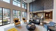 This single-floor community space in Stafford, Texas, is defined by its dramatic double-height ceilings and floor-to-ceiling windows that flood the interior with natural light while offering views of the surrounding outdoor courtyard. The architectural design blends contemporary materials—textured tile accent walls, warm wood paneling, geometric metal partitions, and light stone flooring—to create a cohesive, upscale atmosphere throughout.

The main lounge area anchors the space with a soaring ceiling volume, a bold dark-tiled feature wall housing a large flat-screen television, and a curated seating arrangement of mustard yellow wingback chairs, a charcoal leather sofa, and a sculptural geometric coffee table. A tufted yellow bench adds a vibrant accent near the media wall.

Adjacent to the lounge, a communal dining area features a long wood table topped with a striking multi-bulb pendant chandelier, surrounded by eclectic mixed seating. This zone transitions naturally into a well-appointed kitchenette, which includes a central island with a dark quartz countertop, mosaic tile backsplash, wood-paneled walls, a slat-wood ceiling, stainless steel appliances, and built-in bench seating. The overall ambiance is modern, welcoming, and thoughtfully layered.