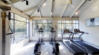 This fitness center is a thoughtfully designed workout space that seamlessly blends architectural elegance with functional amenity. The room is defined by its striking vaulted ceiling with exposed white-painted wood beams, lending a sense of openness and volume that elevates the overall atmosphere. A shiplap ceiling treatment adds a refined, contemporary-rustic character throughout the space.

Natural light pours in abundantly through large, multi-pane windows that wrap the room, framing beautiful views of the surrounding wooded landscape. This connection to the outdoors creates a refreshing and motivating environment for exercise. Track lighting and ceiling-mounted pendant fixtures supplement natural light, ensuring the space is well-illuminated at all times.

The fitness center is outfitted with a selection of cardio equipment, including multiple elliptical machines and a treadmill, all positioned to take full advantage of the scenic outdoor views. The flooring is carpeted, providing comfort underfoot during workouts. A ceiling fan is also present, contributing to air circulation and comfort within the space.

Overall, the fitness center strikes a balance between a high-performance workout environment and a welcoming, architecturally distinctive interior, making it an inviting destination for daily exercise.