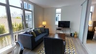This modern single-floor space offers a well-designed and inviting living environment defined by clean lines and a bright, airy atmosphere. The living area is a standout feature, bathed in natural light through large floor-to-ceiling and standard windows that frame views of the surrounding neighborhood. Light-toned walls complement the warm hardwood flooring, creating a cohesive and welcoming aesthetic throughout.

The living room is thoughtfully furnished with a charcoal-gray sofa accented by green and gray throw pillows, paired with a matching armchair, both arranged around a bold geometric-patterned area rug in yellow and white. A flat-screen television sits on a sleek metal and wood console, completing the entertainment setup. A classic table lamp adds a warm ambient glow to balance the abundant natural light.

The overall ambiance of the space is contemporary and functional, with a neutral color palette punctuated by carefully chosen accent pieces that add personality without overwhelming the design. The open layout enhances the sense of spaciousness, making the area feel both comfortable and practical. This space is well-suited for those who appreciate modern design, natural light, and a clean, organized living environment.