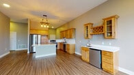 This single-floor unit showcases a thoughtfully designed open-concept layout with wood-tone luxury vinyl plank flooring running throughout the main living and kitchen areas. The kitchen is a standout feature, equipped with warm natural knotty wood cabinetry extending to the ceiling, crisp white countertops, stainless steel appliances including a refrigerator, range, and dishwasher, and a central island with bar-height seating. A built-in wine rack and a secondary wet bar with its own sink and under-counter refrigerator add a refined, functional touch. A rustic pendant light fixture anchors the kitchen, enhancing its character.

The living area connects seamlessly to a balcony through large windows that invite natural light. Bedrooms are finished with plush wall-to-wall carpet, ceiling fans, and vaulted ceilings that add an airy sense of volume. At least one bedroom connects directly to a bathroom, offering added privacy. Bathrooms are appointed with clean white tub-shower combos featuring handheld showerheads and brushed nickel hardware. Neutral gray tones throughout the walls create a cohesive, modern aesthetic across the entire space.
