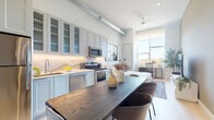This thoughtfully designed studio presents a seamless open-plan layout where the kitchen, dining, and living areas flow naturally into one another. The kitchen is outfitted with soft grey shaker-style cabinetry — including glass-front upper cabinets — paired with a striking marble-look backsplash and countertops, under-cabinet lighting, and a full suite of stainless steel appliances. A substantial dark wood dining table anchors the center of the space, providing generous surface area for both meals and work.

An exposed metallic HVAC duct runs overhead, lending the space an industrial-chic character that contrasts elegantly with the otherwise clean, contemporary aesthetic. Light hardwood flooring runs throughout, contributing to the bright, airy feel of the interior.

The sleeping area sits adjacent to the main living zone, bathed in natural light from oversized floor-to-ceiling windows that offer open city views. Soaring ceiling heights amplify the sense of volume, while a ceiling fan ensures year-round comfort. A dedicated workspace is neatly integrated into the windowed corner.

The bathroom is finished to a high standard, featuring a wall-mounted floating vanity with a white undermount sink, warm gold-accented drawer pulls, a large illuminated mirror, and a full-width glass-enclosed walk-in shower clad in neutral stone-look tile. Recessed lighting throughout the unit enhances the polished, modern ambiance.