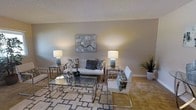 This single-floor space showcases a thoughtfully designed living area that blends contemporary style with a warm, inviting ambiance. The room is anchored by a plush white sofa adorned with decorative pillows in floral and geometric patterns, complemented by a sleek, chrome-framed accent chair with quilted white upholstery. A geometric glass-top coffee table with a chrome base serves as the centerpiece, resting atop a patterned area rug that adds visual texture to the carpeted floor. Symmetrical chrome side tables flank the sofa, each topped with cylindrical table lamps that cast a warm, ambient glow throughout the space. The neutral, taupe-toned walls provide a sophisticated backdrop, enhanced by a large abstract canvas artwork displayed prominently above the sofa. A lush potted plant in the corner introduces a natural element, softening the room's contemporary aesthetic. The textured ceiling adds architectural character, while the overall color palette of whites, grays, and warm beiges creates a cohesive and serene atmosphere. The space is well-proportioned, offering a comfortable and functional layout ideal for relaxed living and entertaining.