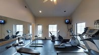 This fitness center offers a thoughtfully designed workout environment built to support a range of exercise routines. The space features soaring vaulted ceilings that create an open, airy atmosphere, complemented by a ceiling fan that helps maintain comfortable airflow during workouts. Natural light streams in through multiple windows, giving the room an inviting and energizing feel. The flooring is covered with dark rubber matting, providing both safety and durability underfoot. A large wall mirror spans one side of the room, enhancing the sense of space while allowing users to monitor their form during exercise. The fitness center is equipped with a selection of cardio machines, including ellipticals and a stationary bike, as well as free weight stations with weight benches and bar racks. A wall-mounted flat-screen television is positioned for easy viewing during workouts. Recessed lighting supplements the natural light, ensuring the space remains well-lit at all times. A door with glass panels provides direct access to the outdoors, further connecting the interior to its surrounding environment. Overall, the space strikes a balance between functionality and comfort, making it a practical and pleasant facility for daily fitness activities.