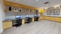 This single-floor business center is designed with functionality and a polished aesthetic in mind. The space is anchored by a long, built-in workstation along one wall, featuring a striking granite countertop paired with warm honey-toned wood cabinetry. Multiple desktop computers are stationed along this surface, each accompanied by ergonomic mesh chairs, making it a practical hub for productivity. Above the workstation, an open display shelf showcases framed artwork and decorative accents, adding a refined touch to the professional environment. A dedicated printing and copying station occupies a separate built-in counter area, flanked by floor-to-ceiling cabinetry for ample storage. Track lighting and recessed ceiling fixtures provide bright, even illumination throughout, complemented by light-toned laminate flooring that lends a clean, contemporary feel underfoot. The open floor plan allows for comfortable circulation throughout the space. A cork bulletin board mounted on one wall serves as a practical information display, while framed artwork adorns the walls to soften the utilitarian atmosphere. Dark-framed interior doors with frosted glass panels add a modern architectural detail. The overall ambiance is professional yet inviting, making this business center a well-equipped and thoughtfully designed space for work and productivity.