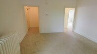 This single-floor space presents a straightforward and functional layout finished in a neutral, light color palette throughout. The main room is carpeted in a beige tone that extends into the adjoining areas, creating a cohesive and understated aesthetic. White walls and white trim provide a clean backdrop, keeping the space feeling bright and open.

Visible from the main room are two doorways leading to additional areas. One opens to a closet or utility space, where a hanging rod is visible, suggesting dedicated storage. The other doorway leads to a secondary room that appears to benefit from natural light, contributing to an airy feel across the unit.

A wall-mounted thermostat or control panel is centrally positioned, indicating climate control functionality. The baseboard trim runs consistently along the walls, adding a simple but polished architectural detail. Overall, the space offers a blank-canvas environment with practical features, suitable for a variety of furnishing arrangements and personal customization.