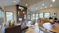 This single-floor space showcases a contemporary mid-century modern design aesthetic, anchored by a striking floor-to-ceiling wood-tiled feature wall that serves as the architectural centerpiece of the room. The feature wall integrates a two-sided fireplace, open display niches, and a wall-mounted television, creating a functional and visually dramatic focal point that divides the lounge and dining areas.

The soaring vaulted ceilings amplify the sense of openness throughout, while amber glass pendant lights suspended at varying heights add warmth and visual interest. The lounge area is furnished with a tufted sectional sofa, mid-century accent chairs, and sculptural wooden side tables arranged over a patterned area rug, contributing to a relaxed yet refined atmosphere.

Adjacent to the lounge, a round dining table with upholstered chairs provides a communal gathering area. Natural stone-look tile flooring extends throughout, complementing the warm wood tones of the built-in cabinetry and kitchenette along one wall, which includes small appliances and a sink. Large windows frame outdoor greenery and pool views, flooding the interior with natural light and reinforcing the connection between indoors and outdoors.