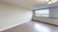 This studio unit at 329 18 Ave SW in Calgary's vibrant Mission neighbourhood offers a well-planned, single-floor layout that maximizes every square foot. The open-concept living area features light-toned laminate hardwood flooring and neutral beige walls, creating a clean, bright atmosphere. A textured ceiling adds subtle visual interest overhead. A large window in the living area frames views of the Calgary skyline, allowing natural light to fill the space while a built-in low-profile cabinet sits beneath the sill, providing additional storage.

The galley-style kitchen is notably well-appointed for a studio, featuring dark wood cabinetry extending to the ceiling for ample storage, a warm metallic backsplash, light quartz countertops with an undermount sink, and a full suite of stainless steel appliances including a refrigerator, stove, and built-in microwave. Track lighting illuminates the workspace effectively.

The bathroom includes a full soaker tub and shower combination with floor-to-ceiling tile surround, a modern vanity with quartz countertop, dark wood cabinetry, and warm accent lighting. The entrance provides a natural transition into the living space, completing this efficiently designed urban studio.