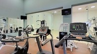This fitness center offers a comprehensive workout environment designed to meet a range of exercise needs. The space is outfitted with an array of Magnum-brand cardio machines, including treadmills, ellipticals, and stationary bikes, providing users with multiple options for cardiovascular training. A multi-functional strength training cable machine is also present, supporting resistance and weight-based workouts. The flooring features a durable, cushioned surface in a warm neutral tone, well-suited for high-traffic fitness use. Natural and artificial lighting combine to create a bright, energizing atmosphere throughout the room. Glass partition walls add a sense of openness and visual connectivity within the space, while wall-mounted flat-screen televisions are positioned for easy viewing during workouts. The light-toned walls and clean design contribute to a modern, professional ambiance. The layout is thoughtfully arranged to allow comfortable movement between equipment, making the fitness center accessible and practical for everyday use.