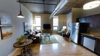 This single-floor loft-style space showcases a compelling blend of industrial design and contemporary comfort. The open layout maximizes the sense of space, with light hardwood flooring running throughout and connecting each functional area seamlessly. Exposed HVAC ductwork runs prominently across the ceiling, lending an authentic industrial aesthetic that defines the overall character of the space.

The living area is generously proportioned, centered around a leather sofa and complementary seating arrangement, including an armchair and accent chair, positioned around a low-profile coffee table atop a patterned area rug. A media console with a flat-screen television serves as the entertainment focal point. Flanking the living area, multiple windows allow natural light to filter in while offering views of surrounding urban architecture, reinforcing the city loft atmosphere.

A compact dining area featuring a bistro-style table with bar stools is positioned near the front, making efficient use of space. Adjacent to this, a stainless steel refrigerator signals the presence of a kitchen area just beyond the frame. A decorative wall clock and framed artwork contribute thoughtful finishing touches throughout.