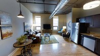 This single-floor loft-style space showcases a compelling blend of industrial design and contemporary comfort. The open layout maximizes the sense of space, with light hardwood flooring running throughout and connecting each functional area seamlessly. Exposed HVAC ductwork runs prominently across the ceiling, lending an authentic industrial aesthetic that defines the overall character of the space.

The living area is generously proportioned, centered around a leather sofa and complementary seating arrangement, including an armchair and accent chair, positioned around a low-profile coffee table atop a patterned area rug. A media console with a flat-screen television serves as the entertainment focal point. Flanking the living area, multiple windows allow natural light to filter in while offering views of surrounding urban architecture, reinforcing the city loft atmosphere.

A compact dining area featuring a bistro-style table with bar stools is positioned near the front, making efficient use of space. Adjacent to this, a stainless steel refrigerator signals the presence of a kitchen area just beyond the frame. A decorative wall clock and framed artwork contribute thoughtful finishing touches throughout.