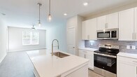 Located at 12400 Dutton Road in Midlothian, VA, this thoughtfully designed one-bedroom, one-bathroom residence offers a clean, modern aesthetic across a single floor. The open-concept layout connects the kitchen and living area seamlessly, creating a bright and functional living space. The kitchen is a standout feature, showcasing crisp white shaker cabinetry, a gray subway tile backsplash, and a full suite of stainless steel appliances including a refrigerator, electric range, and built-in microwave. A central island with a white quartz countertop and an integrated sink is highlighted by stylish pendant lighting, making it ideal for both meal prep and casual dining. Light wood-look flooring flows consistently throughout the unit, complementing the neutral warm-gray walls and white trim. The living area benefits from ample natural light through windows with views of the surrounding greenery. The bedroom is a comfortable retreat with a ceiling fan, a large window, and access to a dedicated closet. The bathroom features a walk-in glass-enclosed shower, a white vanity with quartz countertop, and brushed nickel hardware and grab bars. A hallway connects the key areas efficiently, completing this well-appointed single-floor residence.