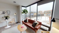 This single-floor residence showcases a thoughtfully designed open-concept layout that balances modern aesthetics with functional living. The living area is a standout, anchored by dramatic floor-to-ceiling corner windows with black frames that flood the space with natural light and offer expansive views of the surrounding urban streetscape. Light wood-toned flooring runs throughout, complementing the neutral white walls and creating a cohesive, airy feel.

The kitchen is fitted with dark wood cabinetry, white countertops, stainless steel appliances including a range, over-the-range microwave, and refrigerator, and a textured three-dimensional tile backsplash that adds visual interest. Storage is efficiently organized along a galley-style layout.

The bedroom is calm and understated, featuring recessed lighting, neutral walls, and soft carpeting, providing a quiet retreat from the open living spaces. The full bathroom presents a clean, contemporary design with a soaking tub and shower combination, light tile flooring, a floating vanity with an integrated sink, and a backlit mirror that adds a refined touch. The overall ambiance throughout the space is modern, bright, and urban.