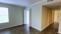 This single-story space presents a clean and understated interior design aesthetic. The main living area is finished with dark hardwood-style flooring that contrasts elegantly against the light gray-green walls, creating a grounded yet airy atmosphere. Crown molding runs along the ceiling perimeter, adding a refined architectural detail that elevates the overall look of the space.

A wide arched opening connects the main room to an adjacent area, suggesting an open and flowing floor plan that promotes a sense of spaciousness. The transition between rooms is marked by a warm beige accent wall, offering a subtle contrast to the cooler tones of the primary living space.

Natural light filters into the room through a glass door fitted with vertical blinds, hinting at outdoor access and contributing to a bright, welcoming ambiance. The white baseboards and door trim provide crisp definition throughout, tying the color palette together cohesively.

The space is move-in ready with a neutral, versatile palette that accommodates a variety of interior design styles, making it an adaptable and functional environment.