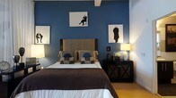 This thoughtfully designed single-floor space makes a strong visual impression through its carefully curated interior aesthetic. The bedroom serves as a standout feature, anchored by a deep navy blue accent wall that provides a rich, dramatic backdrop to the room. A queen-sized bed with a textured, patterned upholstered headboard in warm neutral tones sits at the center, dressed in crisp white linens and layered with decorative pillows in coordinating earth tones and blue circular accents. Flanking the bed are matching dark wood nightstands with an X-frame design, each topped with elegant white ceramic lamps that cast a warm, inviting glow. A curated gallery of framed figurative artwork is displayed across the blue wall, lending the space a gallery-like, artistic character. The warm-toned hardwood flooring adds a natural contrast to the cool blue walls, creating a balanced and grounded atmosphere. Exposed ceiling beams add an architectural detail that enhances the room's character and depth. The overall ambiance of the space is one of refined sophistication, blending bold color choices with classic furnishings to create an environment that feels both stylish and comfortable.