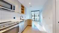 This single-floor unit showcases a thoughtfully designed contemporary interior with a clean, minimalist aesthetic throughout. Light-toned hardwood flooring extends seamlessly across the living and kitchen areas, reflecting abundant natural light and contributing to a bright, airy atmosphere.

The kitchen is well-appointed with a full suite of stainless steel appliances, including a range with oven, built-in microwave, dishwasher, and refrigerator. Warm wood cabinetry is paired with light gray upper cabinets and open shelving, while a white vertical subway tile backsplash adds texture and visual interest. A deep undermount sink with a matte black gooseneck faucet and ample counter space complete the functional layout.

A floor-to-ceiling black-framed window anchors the living area, drawing in generous daylight and offering views of the surrounding urban streetscape. The open layout creates a sense of spaciousness, with crisp white walls providing a neutral, versatile backdrop.

The bathroom features dark mosaic tile flooring, a wall-mounted illuminated mirror, a wood-toned vanity with a white countertop, and matte black hardware and grab bars, combining practicality with a refined modern finish.

Overall, the space presents a cohesive, contemporary design with high-quality finishes, ample natural light, and a well-organized layout suited for comfortable everyday living.