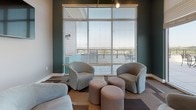 Located at 910 Goddin St in Richmond, Virginia, this single-floor amenity lounge is a thoughtfully designed communal space that blends contemporary style with functional comfort. The open floor plan seamlessly transitions between distinct zones, each serving a unique purpose while maintaining a cohesive aesthetic throughout.

The lounge area is anchored by plush, rounded barrel chairs and soft ottomans arranged on a geometric patterned carpet, creating an inviting setting for relaxed conversation or casual gatherings. A wall-mounted flat-screen television adds a media-ready element to this comfortable retreat. Deep teal accent walls contrast elegantly with light grey and white tones, lending the space a modern, sophisticated character.

Large floor-to-ceiling windows and wide sliding glass doors flood the interior with abundant natural light and frame sweeping views of the surrounding Richmond skyline and greenery. These doors open directly onto a spacious outdoor terrace furnished with dining sets and umbrellas, making for a seamless indoor-outdoor experience.

The kitchenette area features warm hardwood flooring, dark cabinetry with granite-style countertops, open shelving, and a full-size stainless steel refrigerator — well-equipped for entertaining. Track lighting and pendant fixtures throughout the space provide warm, layered illumination that complements the overall inviting ambiance.