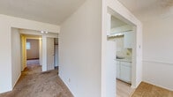 This single-floor unit offers a straightforward, functional layout connecting its primary living spaces through open archways and a central hallway. The kitchen is efficiently designed with white cabinetry, white appliances including a refrigerator, electric range, and built-in oven, along with a single-basin sink and light-toned countertops. Hard-surface flooring runs through the kitchen, transitioning to wall-to-wall carpet in the living and bedroom areas, which lends warmth throughout. The living area benefits from a ceiling fan with integrated lighting and a window fitted with horizontal blinds, allowing natural light into the space. Bedrooms are carpeted and feature ceiling fans, windows with blinds, and sliding closet doors that open to reveal rod-and-shelf storage systems. Walls throughout carry a neutral, cream-toned palette, complemented by textured ceilings. The bathroom is equipped with a full-size soaking tub surrounded by tiled walls, a towel bar, and a vanity with a white countertop, set on wood-look hard flooring. Overall, the unit maintains a clean, neutral aesthetic suitable for comfortable, everyday living.